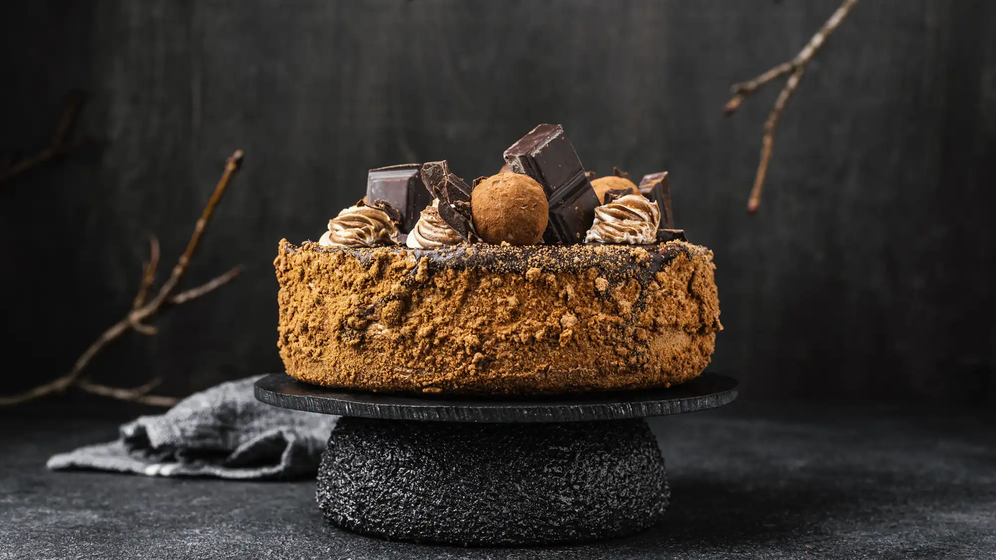Bogato dekorisana čokoladna torta sa komadićima čokolade, čokoladnim kuglicama i kremom, postavljena na elegantnom crnom postolju.