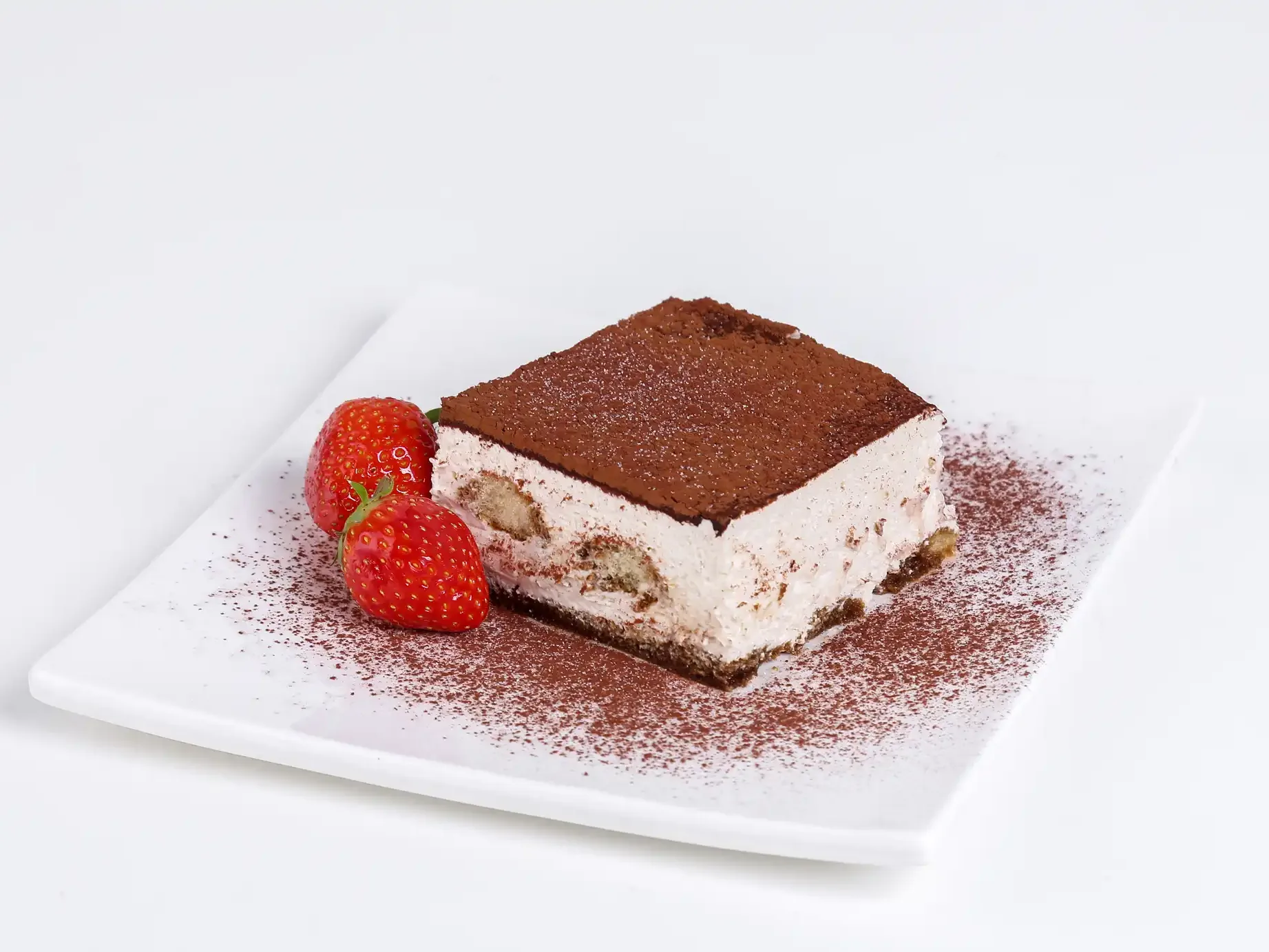 Komad kolača tiramisu na bijelom tanjiru, posut kakaom, sa dvije svježe jagode.