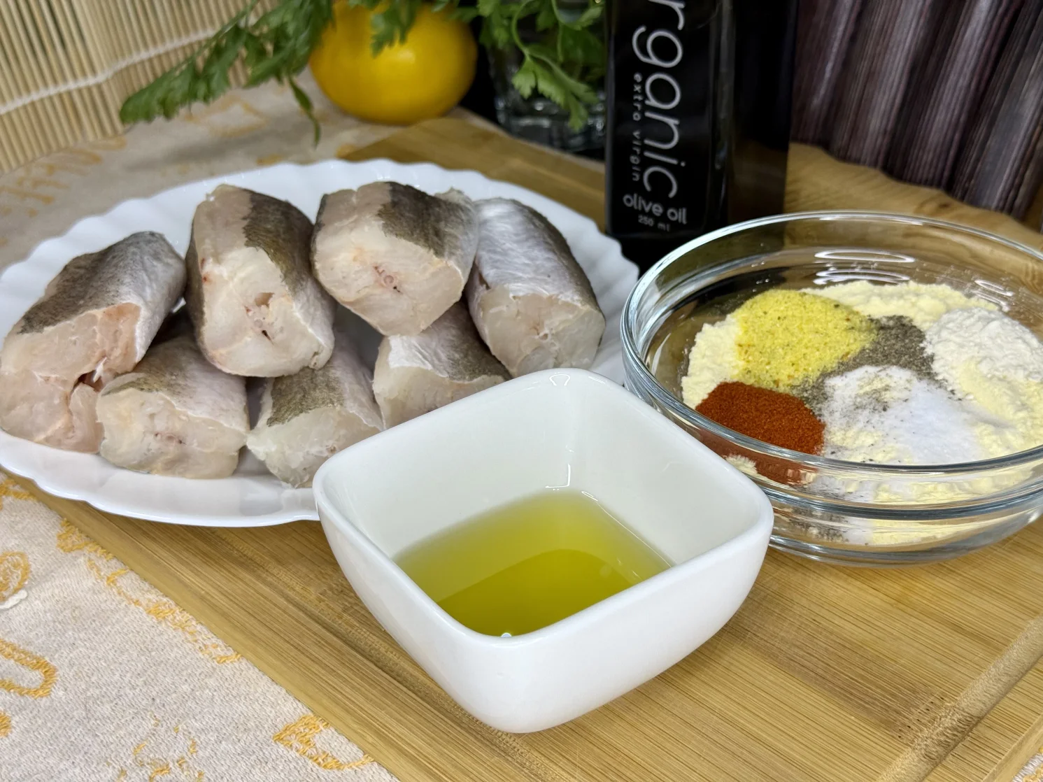 Očišćeni komadi oslića na tanjiru, u zdjeli kukuruzno brašno, so, biber, aleva paprika, bijeli luk u prahu, pored činija sa uljem. Sve spremno za pečenje pohovanog oslića u rerni.